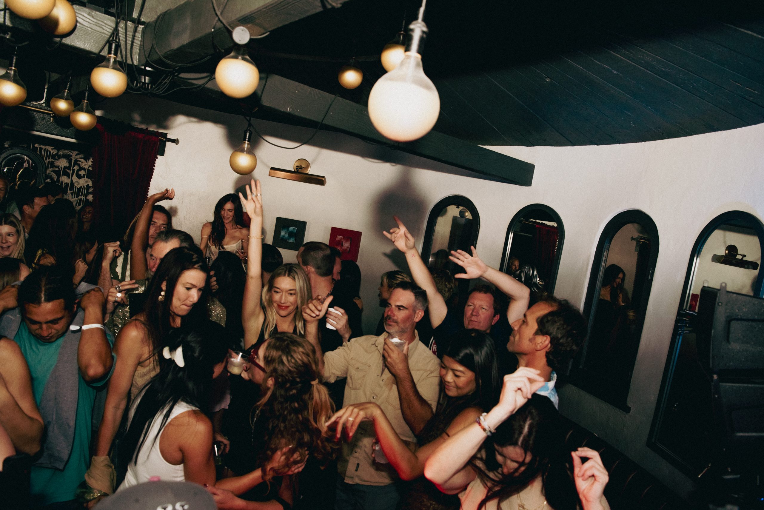 Guests dancing with their hands raised on a packed indoor dance floor beneath hanging globe lights.