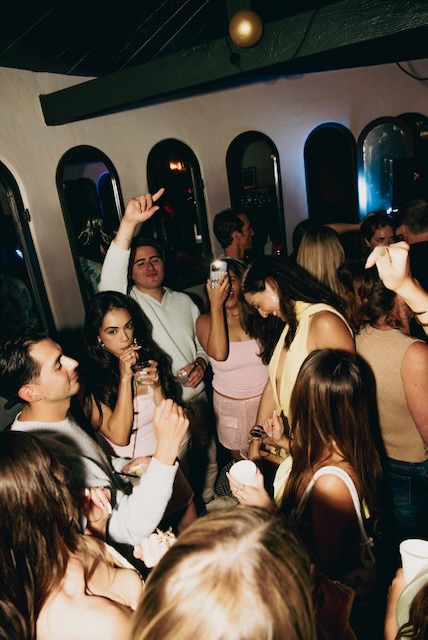 Guests holding drinks and dancing beside a row of arched wall mirrors.