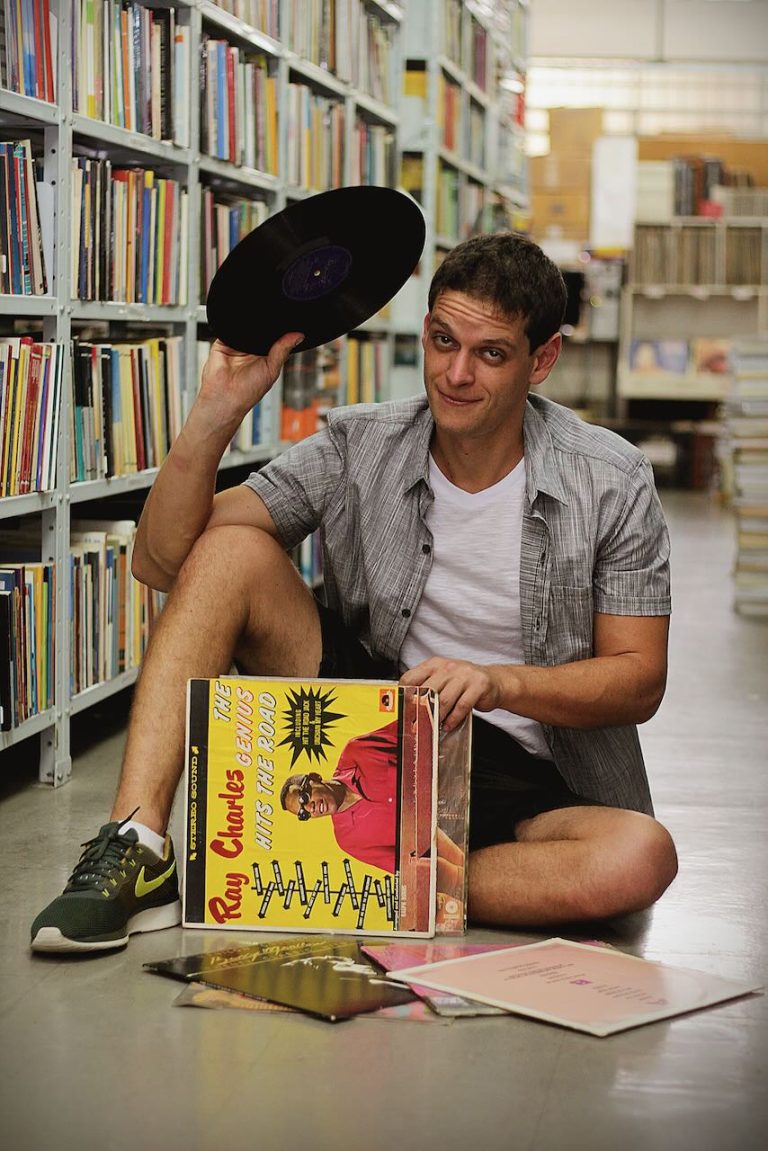 Caiio seated between record shelves, holding a vinyl record beside a stack of albums.