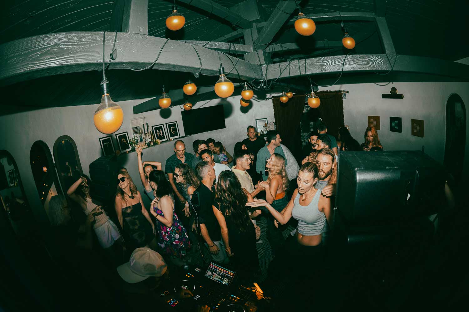 Guests dance close to a DJ booth beneath warm globe lights in a room with exposed beams.