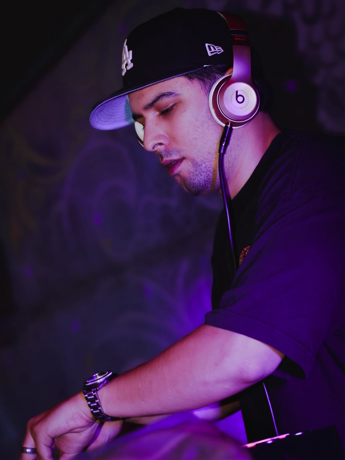 DJ wearing a black LA cap and Beats headphones performs under purple club lighting, focused on the controls in front of him.