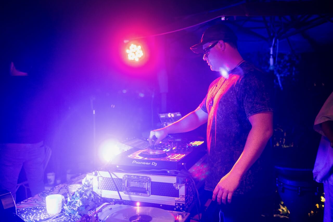 DJ mixing on a Pioneer DJ controller under vivid purple, blue, and magenta lighting at a nighttime event.