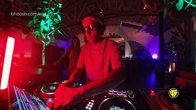 DJ wearing sunglasses and headphones performing at the decks under intense red and purple lighting.