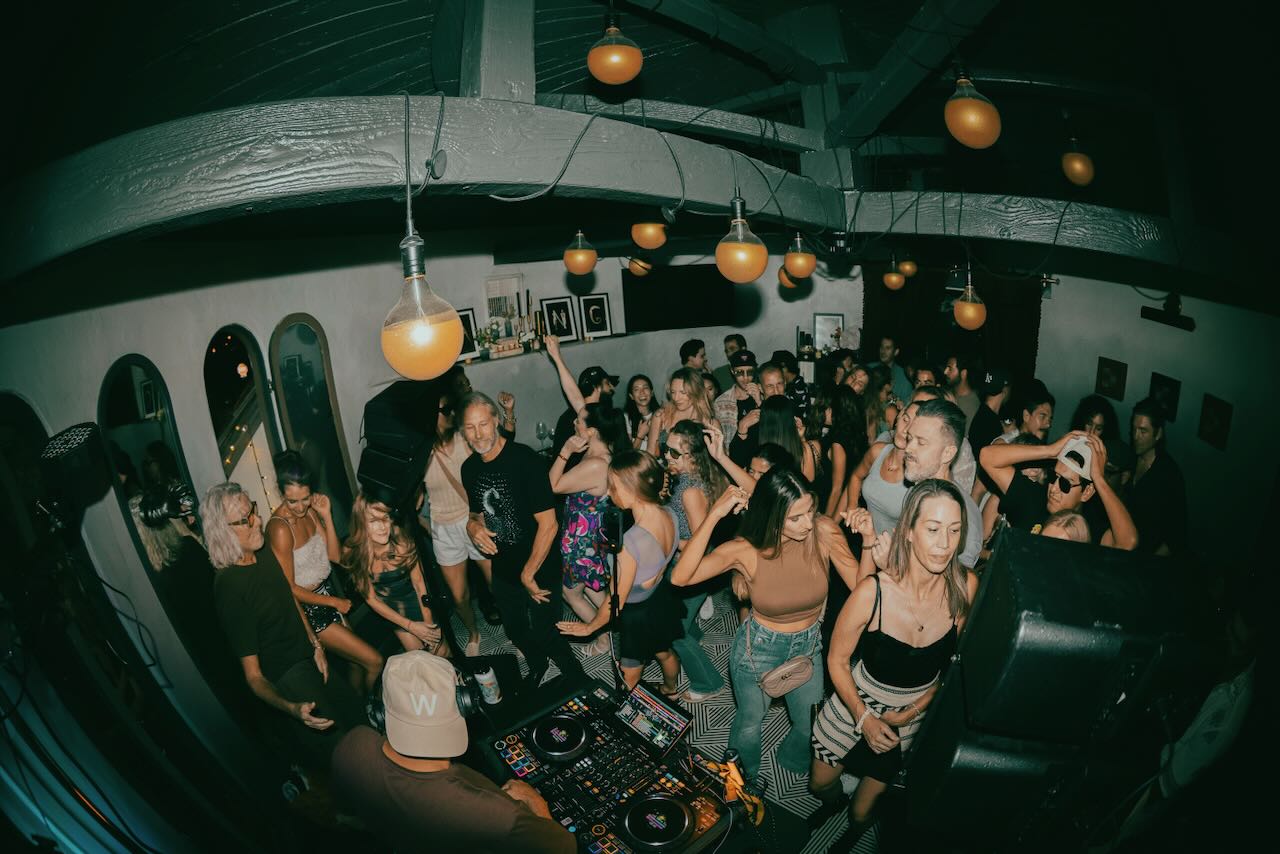 A crowd dances around the DJ booth beneath globe lights and exposed ceiling beams.