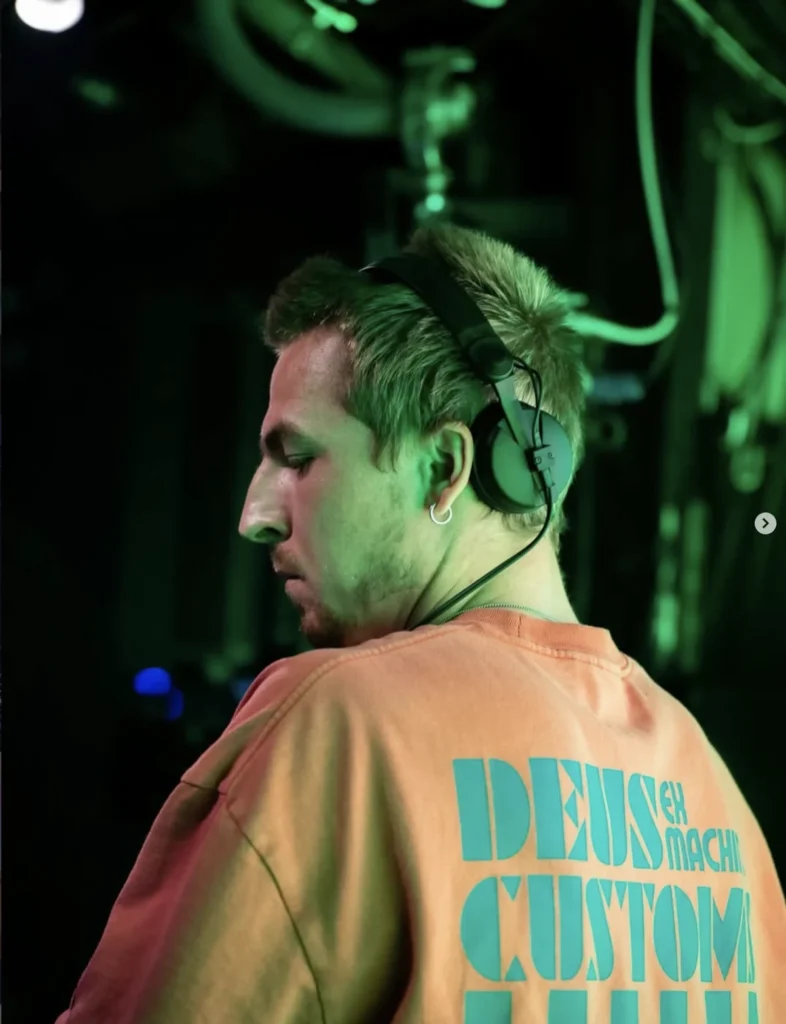 James Blonde wearing headphones at a DJ booth under green lighting.