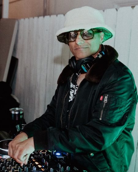 A DJ wearing a white bucket hat, sunglasses, and headphones around his neck adjusts the mixer at a nighttime event, lit by green stage lighting.